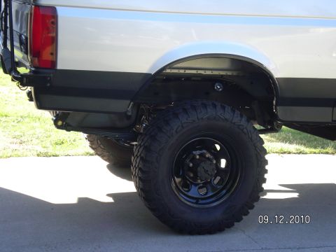 1996 Ford Bronco - Rear quarter of a lifted silver full-size SUV on a driveway, showing large mud-terrain tires on black steel eight-lug wheels and a big gap…