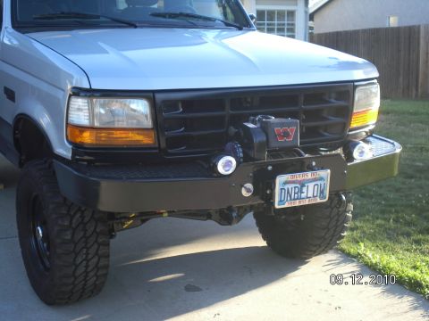 1996 Ford Bronco - Three-quarter front view of a lifted white 1990s Ford SUV on a driveway, wearing a black diamond-plate winch bumper with a Warn winch, roun…