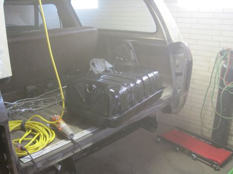 1979 Ford Bronco - Freshly black-coated metal fuel tank sitting on a vehicle frame in a garage, with yellow air hose and red creeper nearby.