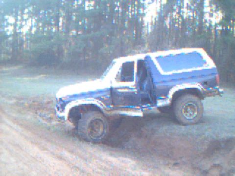 1986 Ford Bronco - This Ford Bronco is parked off-road, with its driver's side door ajar, surrounded by trees.