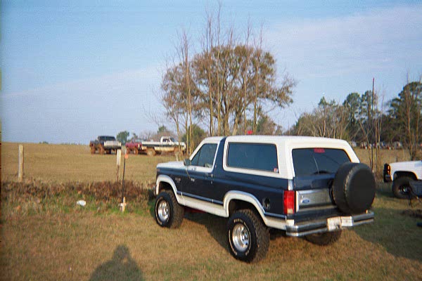 1986 Ford Bronco, blue and white, with spare tire mounted on rear door. Other trucks visible in background