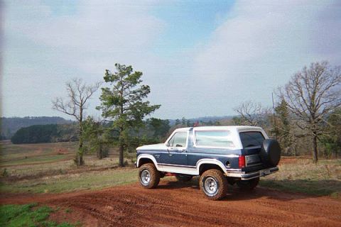 1986 Ford Bronco - A blue and white Ford Bronco is parked on a dirt mound with a wooded landscape behind it.