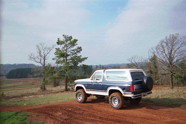 Blue and white Ford Bronco parked on a dirt hill, trees and fields in background