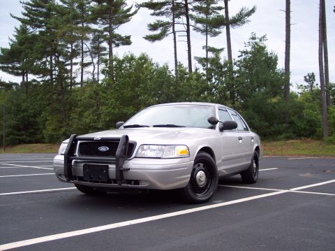 2001 Ford Crown Victoria - A silver 2001 Ford Crown Victoria police car is parked in an empty lot with trees in the background.