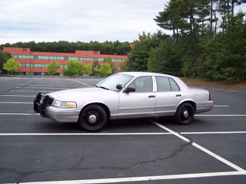 2001 Ford Crown Victoria - A silver 2001 Ford Crown Victoria police car is parked in a lot with a building and trees in the background.