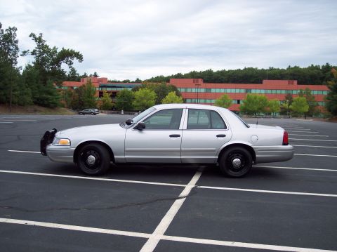 2001 Ford Crown Victoria - A silver 2001 Ford Crown Victoria police car is parked in a lot with a large brick building behind it.