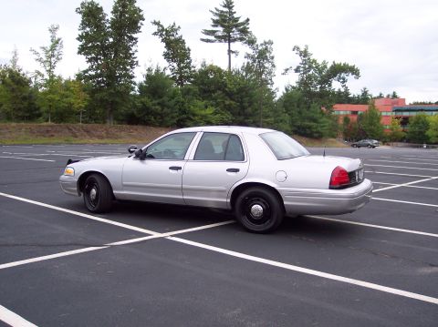 2001 Ford Crown Victoria - A silver police car is parked in a lot with trees and a building in the background.