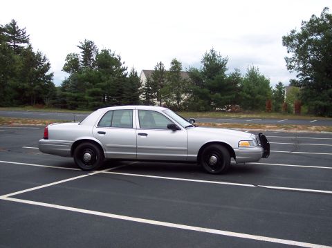 2001 Ford Crown Victoria - A silver 2001 Ford Crown Victoria police car is parked in a lot with trees in the background.
