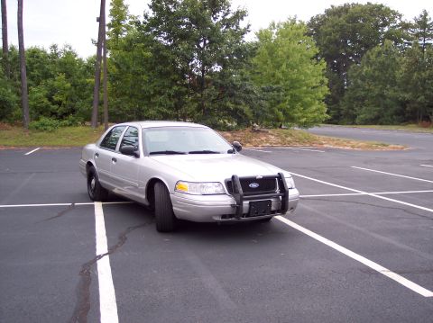 2001 Ford Crown Victoria - A silver 2001 Ford Crown Victoria police car is parked in a lot with trees in the background.