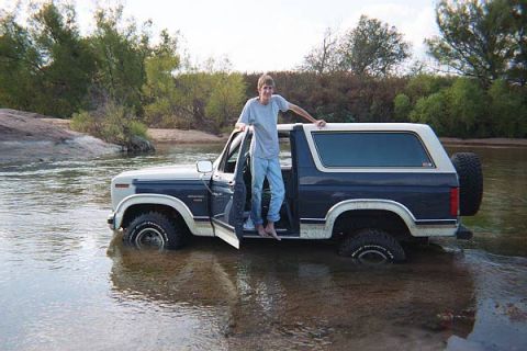 1986 Ford Bronco - A person stands in the open driver's door of a blue and white 1986 Ford Bronco submerged in a river.