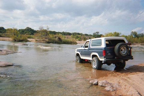 1986 Ford Bronco - 1986 Ford Bronco navigates shallow river crossing with trees and cloudy sky in background.