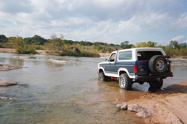Blue and white Ford Bronco with rear spare carrier entering a shallow rocky river, rear three-quarter view