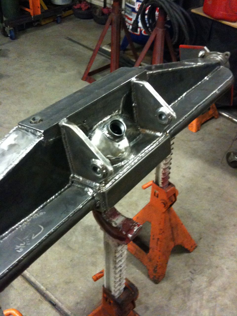 Fabricated metal axle housing with welded brackets, supported by orange jack stands in a workshop