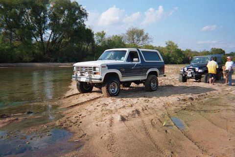 1986 Ford Bronco - A two-tone blue and white Ford Bronco is parked on a sandy riverbank next to a dark-colored Jeep.