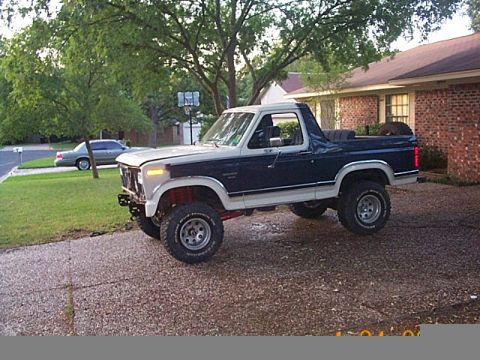 1986 Ford Bronco - A lifted two-tone Ford Bronco sits parked on a gravel driveway in front of a brick house.