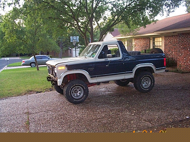 Lifted blue and white 1980s Ford Bronco with top removed, parked in a driveway in front of a brick house