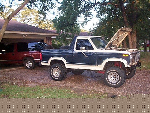 Blue-and-white lifted Ford Bronco, side view with hood up in gravel driveway