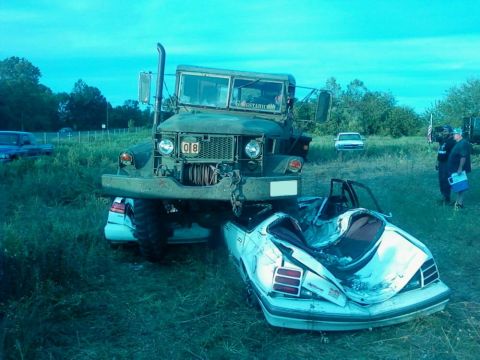 1990 Ford Bronco - A military truck has crushed a white car in a field. The truck's front is visible, and the car is severely damaged.