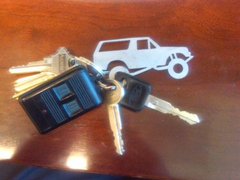 1986 Ford Bronco - A set of keys rests on a wooden surface, accompanied by a metal keychain shaped like a Ford Bronco.