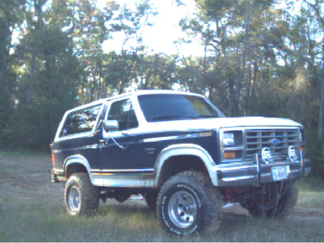 Lifted dark blue and white Ford Bronco with grille guard and white-letter tires parked by trees