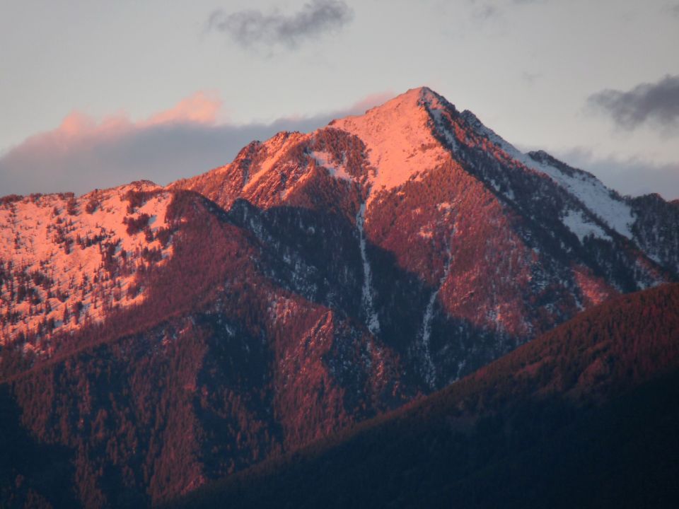 Mountain peaks lit by sunset, with snow patches and dark forests