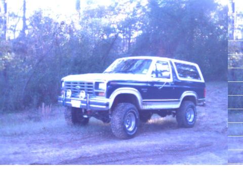 1986 Ford Bronco - This Ford Bronco is parked off-road with trees in the background. It has a two-tone paint scheme and auxiliary lights.