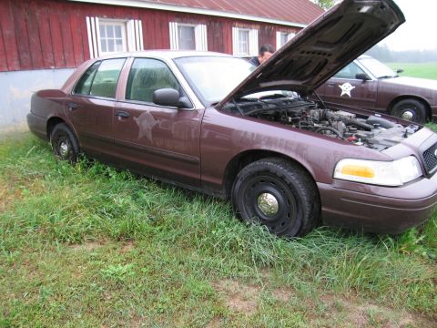 1991 Ford LTD Crown Victoria - Two Ford Crown Victoria police cars are parked in the grass near a red barn, one with its hood open.
