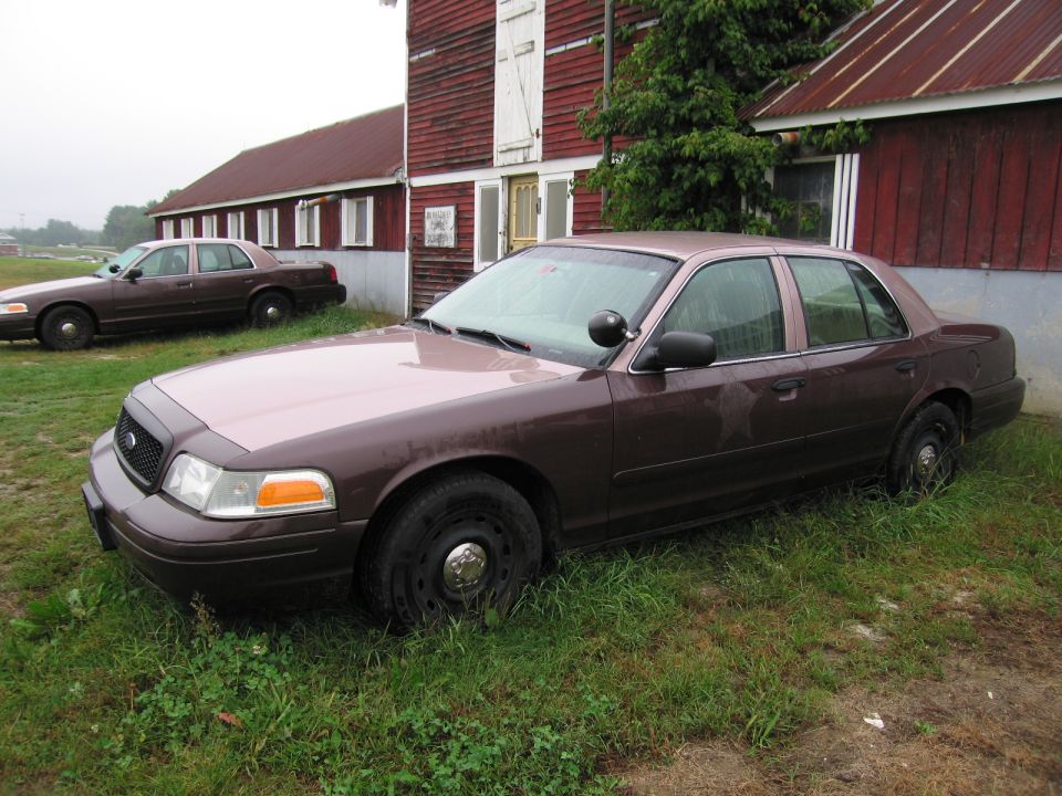 Brown Ford Crown Victoria parked on grass in front of a red barn, another identical car behind it