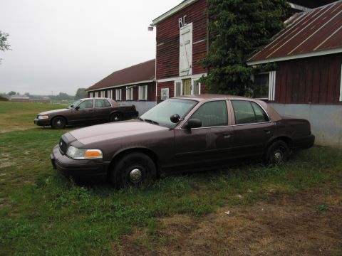 1991 Ford LTD Crown Victoria - Two Ford Crown Victoria Police Interceptors are parked on the grass next to a red barn on an overcast day.