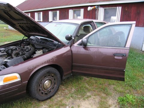 1991 Ford LTD Crown Victoria - Brown Ford Crown Victoria police car with hood and driver door open, parked near a red building.