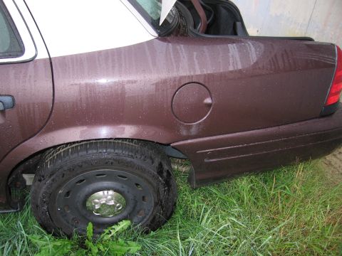 1991 Ford LTD Crown Victoria - A brown sedan with a white roof is parked in the grass, showing its rear quarter panel and a flat tire.