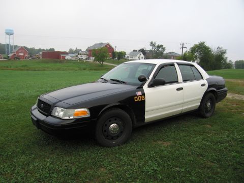 1991 Ford LTD Crown Victoria - A black and white police car, possibly a Ford Crown Victoria, is parked in a grassy field with farm buildings in the background.