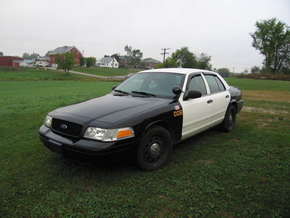 Black and white police car with number 006 on the door, parked on grass with houses in background