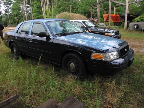 1991 Ford LTD Crown Victoria - Two police cars are parked in a grassy area with some industrial equipment in the background.