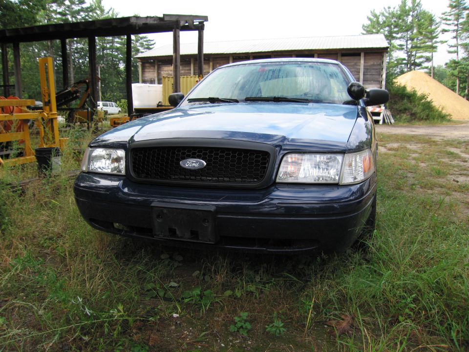 Front view of a dark blue Ford Crown Victoria parked in a grassy area with a wooden structure and a pile of material in the background