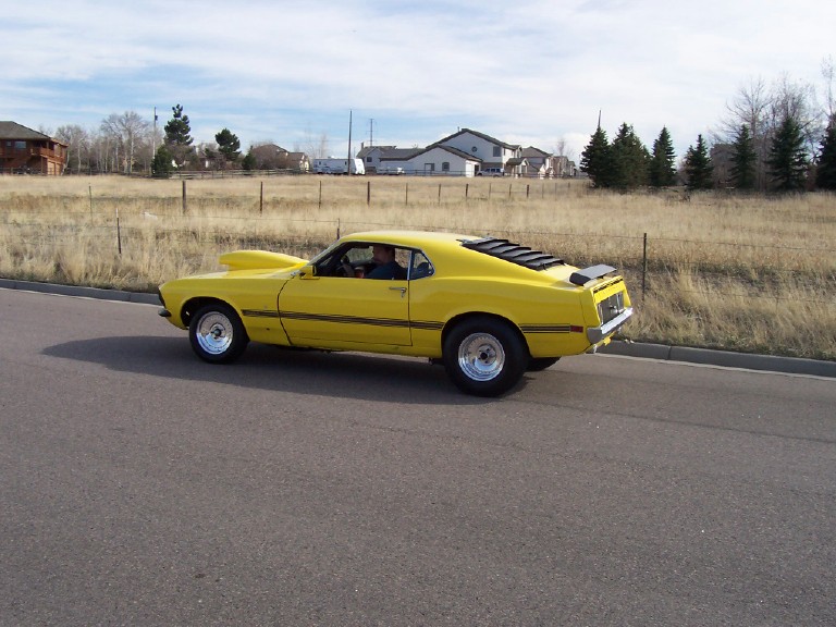 Yellow muscle car with louvers on rear window and a hood scoop, driving on a road
