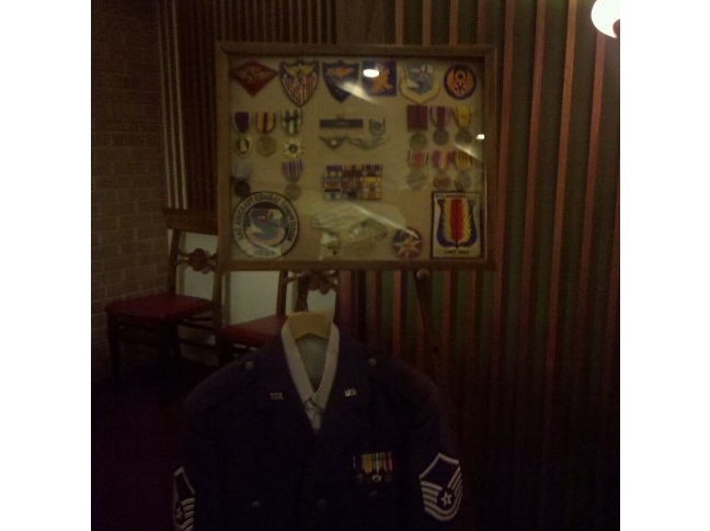 Framed military medals and patches above a dress uniform jacket on a wood-paneled wall