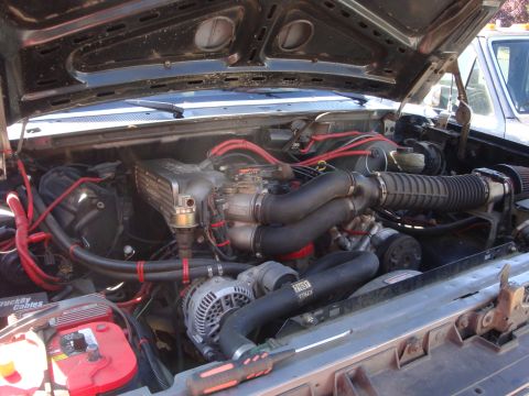 1989 Ford Bronco - Engine bay view of a V8 engine, with parts like the alternator and battery visible. Another vehicle is in the background.