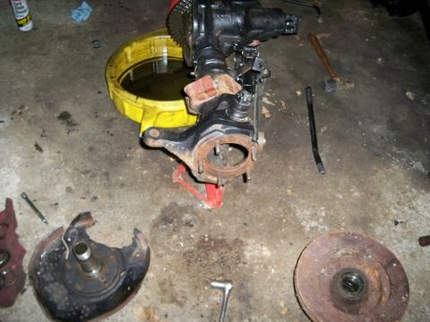 1995 Ford F-150 - A disassembled front axle assembly sits on a jack next to a drain pan and tools, with a broken brake rotor nearby.