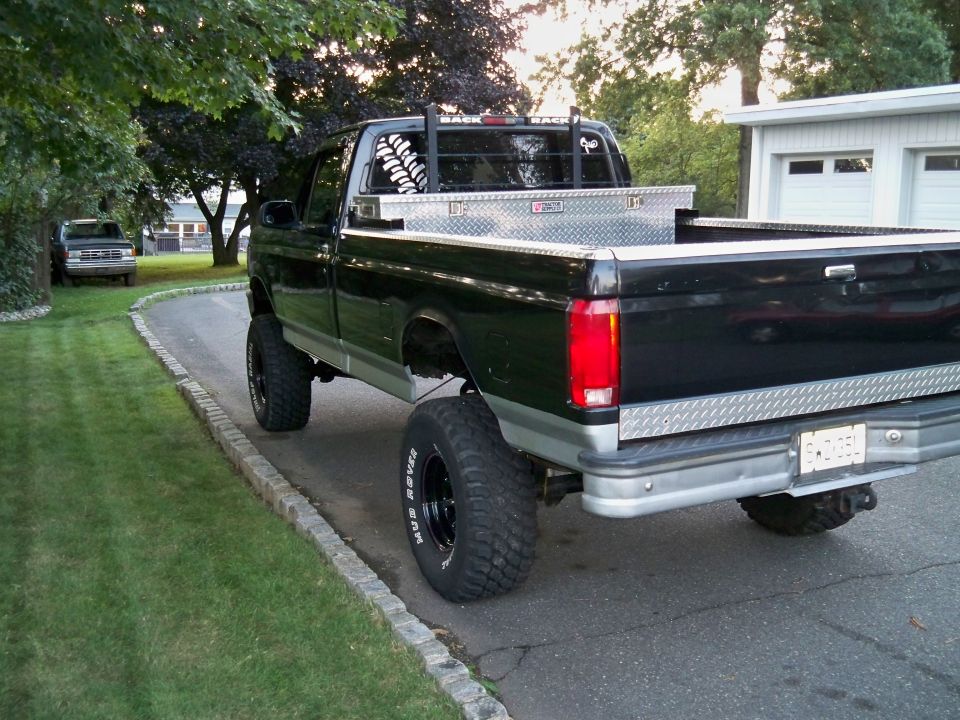 Black pickup truck with lifted suspension and large tires, parked on a driveway. A second truck is visible in the background