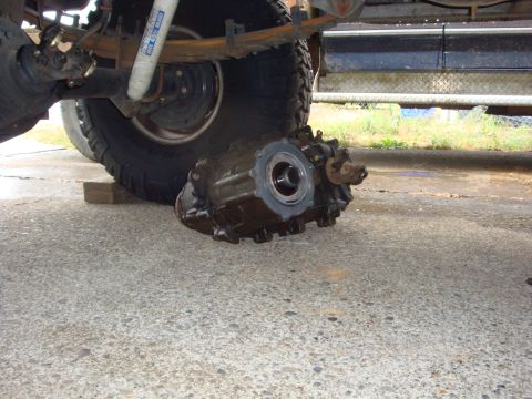 1989 Ford Bronco - Transfer case removed from a vehicle, resting on concrete near the rear tire and suspension.