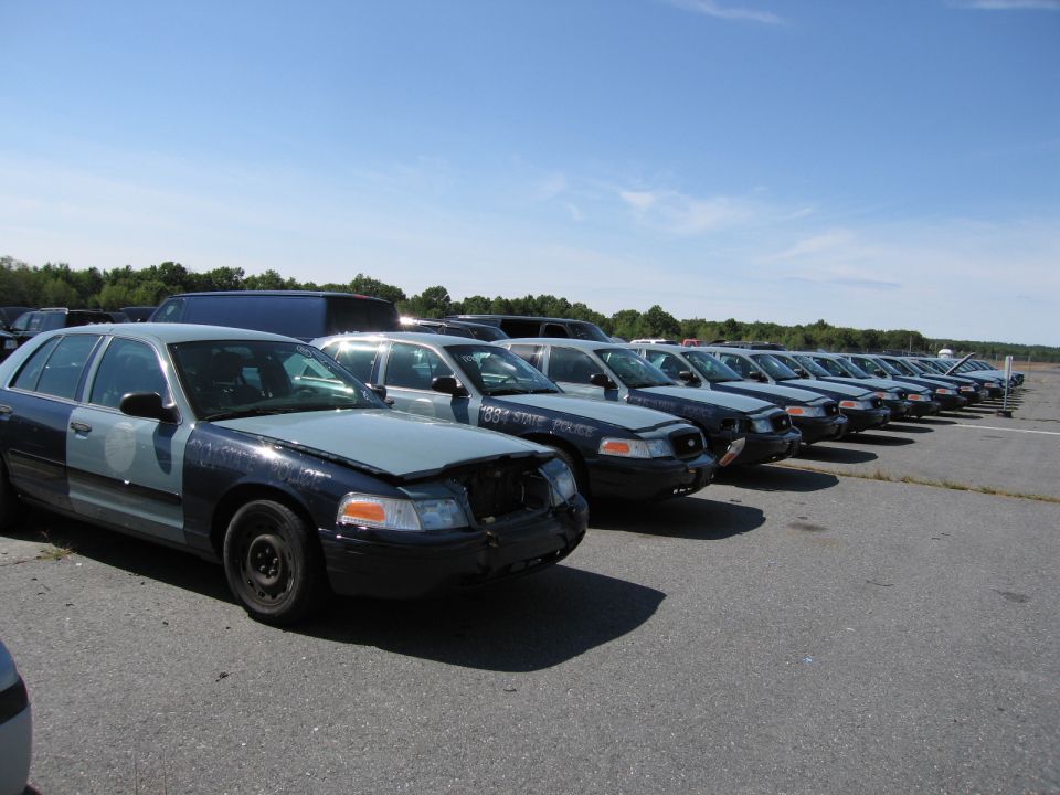 Line of dark blue and light blue Ford Crown Victoria police cars, one with hood open, parked in a lot