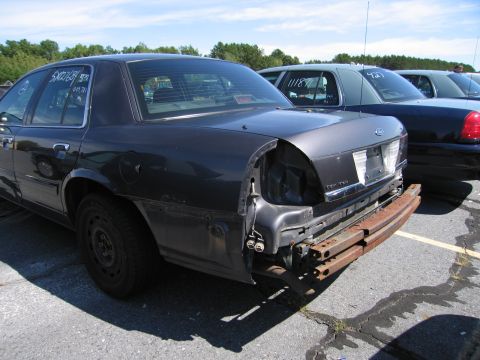 1991 Ford LTD Crown Victoria - A dark sedan is seen from the rear, missing its bumper and one taillight, with other vehicles parked behind it.