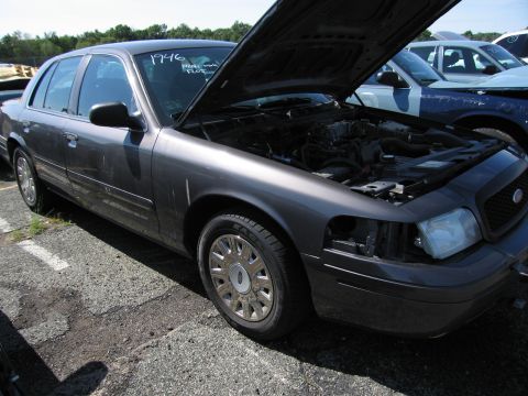 1991 Ford LTD Crown Victoria - A dark gray Ford Crown Victoria sits in a junkyard with its hood open and a headlight missing.