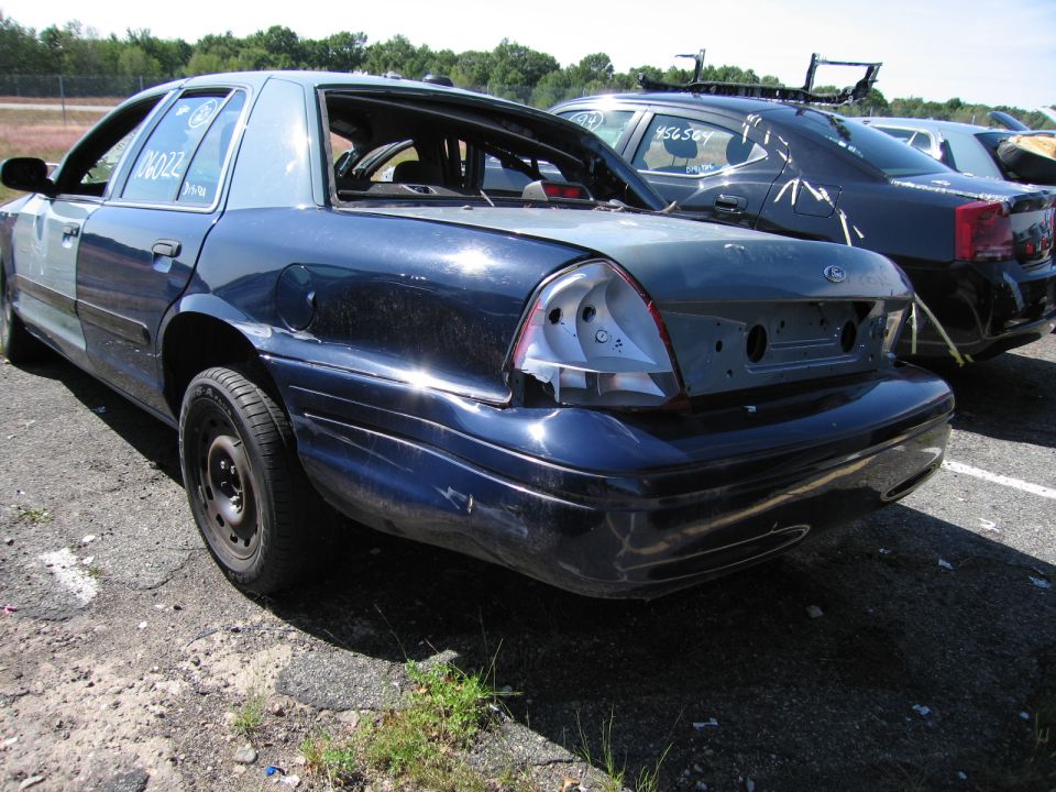 Rear of a dark blue Ford Crown Victoria with missing taillights and rear bumper. Another car is parked behind it