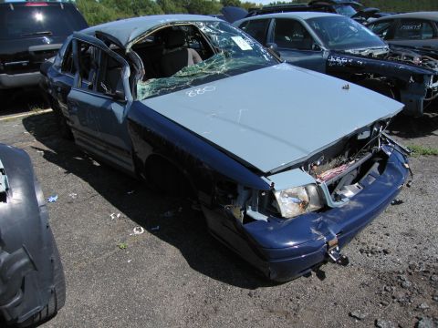 1991 Ford LTD Crown Victoria - This heavily damaged sedan, possibly a Ford Crown Victoria, sits in a junkyard with its windshield cracked and driver's door ajar.