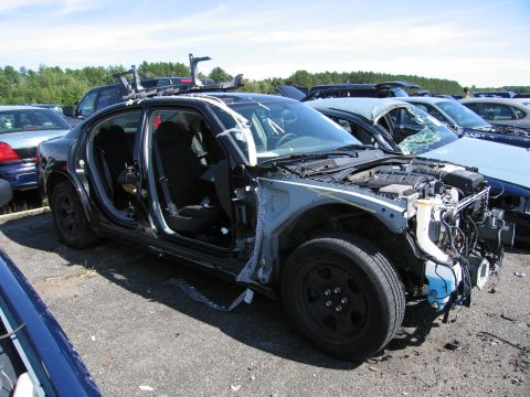 1991 Ford LTD Crown Victoria - A heavily damaged black Dodge Charger is parked in a junkyard with its doors open and front end dismantled.