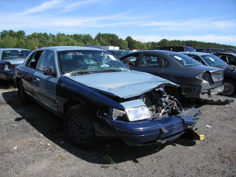 1991 Ford LTD Crown Victoria - A severely damaged sedan sits in a junkyard with other vehicles visible in the background under a clear sky.