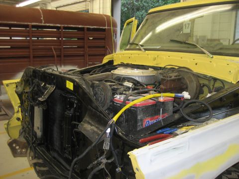 1978 Ford Bronco - The engine bay of a yellow truck is visible with the hood open, revealing the engine and battery. A large brown trailer is parked behind it.
