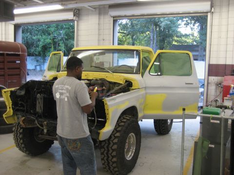 1978 Ford Bronco - A mechanic works on a yellow and grey 1978 Ford Bronco in a garage, with its doors open and hood raised.